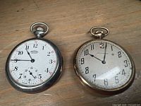 Photo of two pocket watches face up on wooden surface showing Ingersoll Junior with white dial, black numerals, and a sub-second dial, and Standard Watch Co. with cream dial and black numerals.