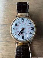 Front view of watch face showing white dial with black Arabic numerals, red 24-hour numerals, black hour and minute hands, red seconds hand