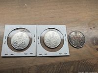 Photo showing the reverse side of two Canadian silver half dollar coins (1964 and 1965) in holders and one loose coin (1966) with the heraldic crest design.
