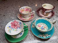Top view of all four cups and saucers showing various floral patterns and colors including pink roses, turquoise, and purple designs.