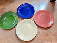 Top view of four round ceramic charger plates in green, blue, coral, and cream colors arranged side by side showing their glazed surface and concentric ring design.