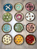 Top angle showing all twelve Florentine gilt coasters arranged in two rows of six, displaying various colors and designs with scalloped gilt edges.
