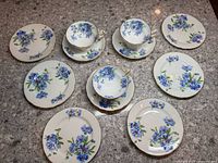 Top-down view of 3 cups on saucers and 6 plates with blue cornflower floral design and gold trim on a granite countertop.