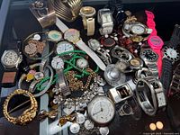 Top-down view of miscellaneous wristwatches, necklaces, brooches, and bracelets arranged on a black surface.