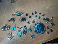 All jewellery pieces arranged on a wooden surface, featuring brooches, pendants, and various stud earrings with iridescent blue-green mother of pearl.