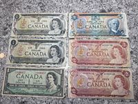 Seven Canadian banknotes laid out on a countertop including three one dollar bills (one from 1954 and two from 1974), two two dollar bills from 1974, and two five dollar bills including one from 1979.