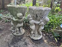 Pair of concrete urn planters with cherubs standing on a base outdoors with surrounding garden vegetation.