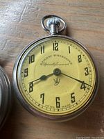 Close-up of JWC Railway Timekeeper pocket watch with yellow dial, black numerals and hands, metal case.