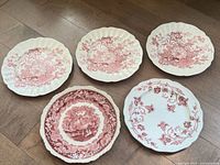 Top-down view of five transferware plates laid out on floor, showing floral and scenic pink/red designs