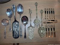 Full lot overview showing silver plate ladles, ornate brass serving pieces, and black-handled brass flatware with Buddha reliefs