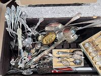 Overview of assorted silverware including serving forks, spoons, collector spoons, and other pieces inside a box.