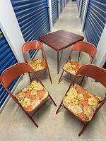 Card table and four folding chairs set seen under overhead lighting, showing orange/red metal frames and floral vinyl padded seats on chairs.
