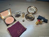 Square Coty compact with powder and mirror, small ornate glass perfume bottle, combined perfume and compact mirror, round Stratton compact, Dorothy Gray lipstick cones in worn black pouch, and burgundy felt Brigham pouch.