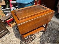 Wooden tea cart with closed double drop leaves, drawer visible, large spoked wheels, small caster wheels missing rubber, and glass serving tray on top.
