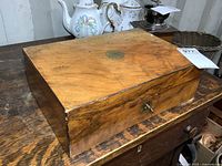 Closed antique wooden writing box sitting on a wooden surface, showing top and side with brass inlay and lock with key inserted.