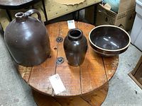 Three items on wooden table: large brown jug, small brown crock, and brown mixing bowl with lighter rim.