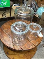 Three clear glass apothecary jars on a round wooden table, all with lids and knob handles, varying in size from small to large.