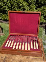 Open wooden case showing 24-piece fish knife and fork set with bone or ivory handles and sterling silver collars, arranged on red velvet lining.