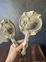 Photo showing front of silver-plated hairbrush and hand mirror with floral Art Nouveau design held against a dark background.