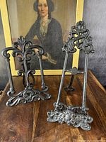 Two antique cast iron easel display stands positioned side-by-side with a portrait in the background, showing overall design and size comparison.