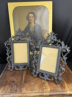 Pair of antique cast iron easel display stands in front of a painted portrait. Both stands feature ornate decorative scrollwork and leaf designs, showing front and angled views.