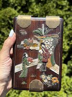 Rectangular hardwood box held in hand outdoors showing carved top surface with three figures and stone inlays in natural scene including flowers and foliage. Brass corner protectors visible.