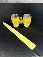 Pair of antique opera glasses with cream barrels, gold rims, central knob, and two matching letter openers with cream handles and yellowish blades on a black surface.