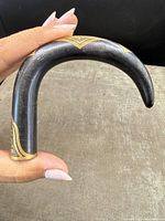 Curved cane handle held in hand showing the blackened metal body with gold inlay design near the curved end.