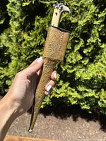 Hand holding an antique Middle Eastern dagger sheathed in an embossed brass scabbard, showing the overall item.