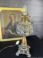 Full view of the antique pierced brass lamp with electric bulb attached, set against a background with a framed painting.