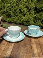 Two Aynsley porcelain teacup and saucer sets shown side-by-side on wooden table outdoors with greenery in background. One set has solid blue teacup; the other has floral decoration inside the cup.