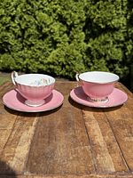 Pair of pink Aynsley teacups and saucers displayed on wooden surface outside, showing both the interior floral pattern and the exterior pink finish.