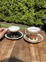Two antique porcelain teacups and matching saucers on a wooden surface outdoors, showing detailed gold accents and patterns on both the Aynsley and Royal Albert sets.