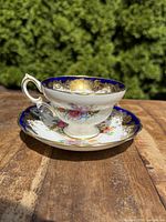 Teacup and saucer set placed on a wooden surface outdoors showing side profile of cup and saucer with sunlight highlighting gold and floral patterns.