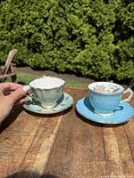 Photo showing two antique porcelain teacups and their matching saucers on a wooden table outdoors, highlighting the floral and gold detailing on both sets.