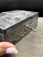 Close-up side angle showing detailed embossed design on silver-metal jewelry box and original brass key inserted in lock.
