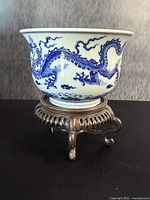 Blue and white porcelain bowl showing dragon design on side, mounted on carved wooden stand with three legs.
