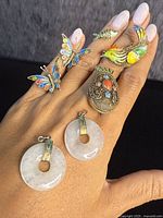 Photo showing six antique Chinese jewelry pieces on hand: two jade pendant earrings, two butterfly enamel rings, bird enamel ring, fish enamel ring, and oval filigree pendant with coral and enamel details.