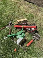Collection of seven antique rustic farm-style kitchen gadgets displayed on grass, including a meat grinder, apple peeler, wire herb chopper, pruners, and various hand tools