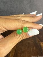 Front view of both green jade stud earrings held between fingers against a neutral background.