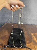 Hand holding the full gold-tone chain necklace with natural white and dark colored pearls against a black pouch on a wooden surface.