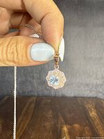 Close-up of pendant held between fingers showing front with blue center stone surrounded by clear stones and rose-gold overlay details.