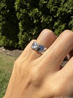 Ring on hand showing side view of clear stone set in wide sterling silver band with polished finish, outdoors with green background.