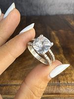 Close-up photo of the sterling silver double-band engagement-style ring held between fingers, showing the large clear round-cut stone and small stones on bands.