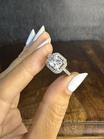 Close-up of ring held between fingers showing the large clear square-cut center stone with halo setting and stone-adorned band.