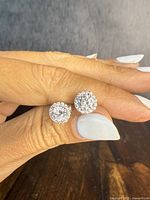 Front view of clear stone halo stud earrings held between fingers showing round stone cluster design.