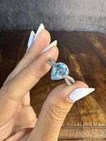 Hand holding the ring showing pear-shaped blue central stone and clear stone halo against a wooden background.