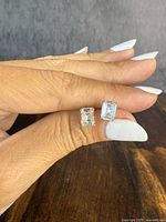 Two emerald-cut clear stone stud earrings held between fingers showing front view, polished sterling silver setting with prong mounts.