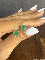 Close-up front view of a pair of pear-shaped green stone sterling silver stud earrings shown on fingers with white nail polish against a wood surface.