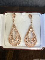 Close-up photo of earring pair in a white cushioned jewelry box showing detailed filigree rose-gold overlay and multiple clear stones on each teardrop earring.